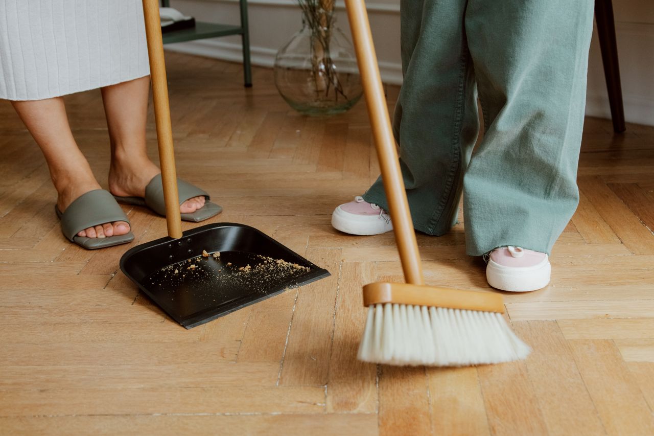 Close-up of people cleaning wooden floor with broom and dustpan indoors.